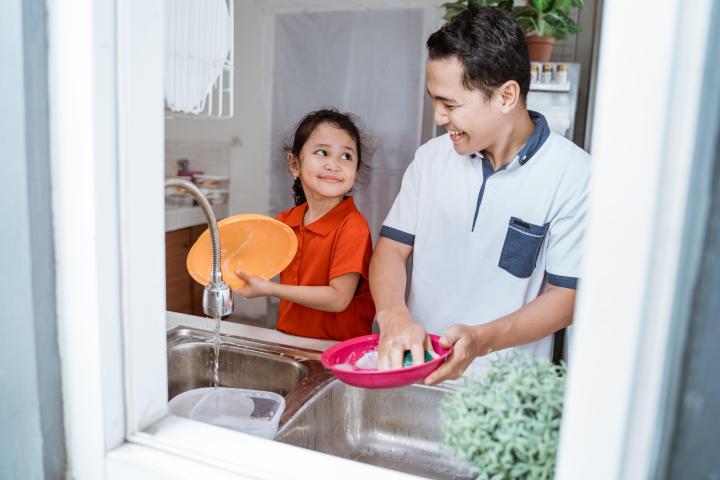 Father and his young daughter together at kitchen sink, washing dishes.