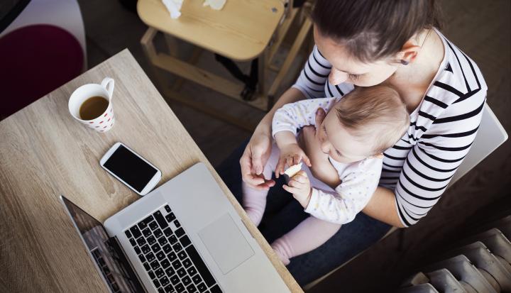 An image showing a woman holding a baby while sitting at a table that holds a laptop, mobile phone and cup of coffee.