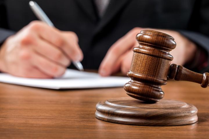 An image showing a gavel resting in the forefront of the picture, while a set of hands writes on a piece of paper in the background. 