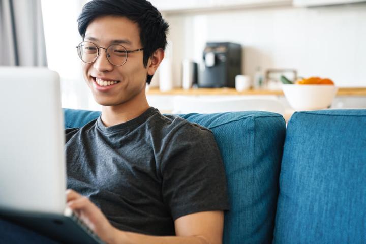 A photo of a man sitting on a couch and smiling while using a laptop.