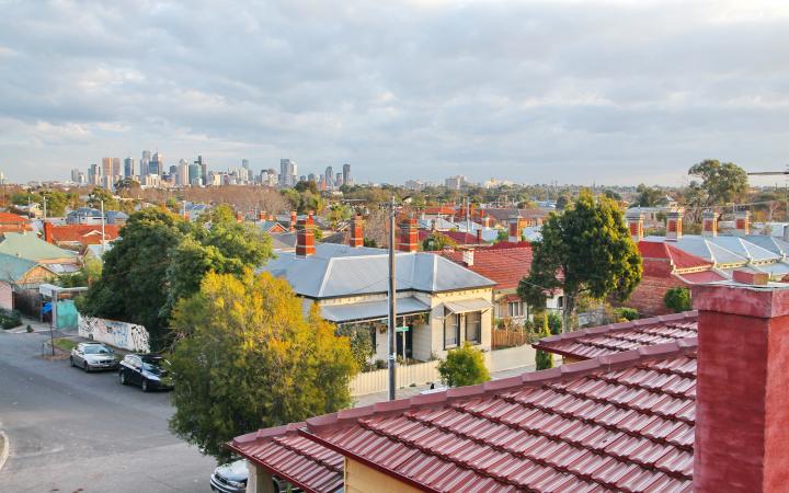 Image showing Melbourne on the horizon, with a suburb sprawling before it. A red roof is in the foreground. 